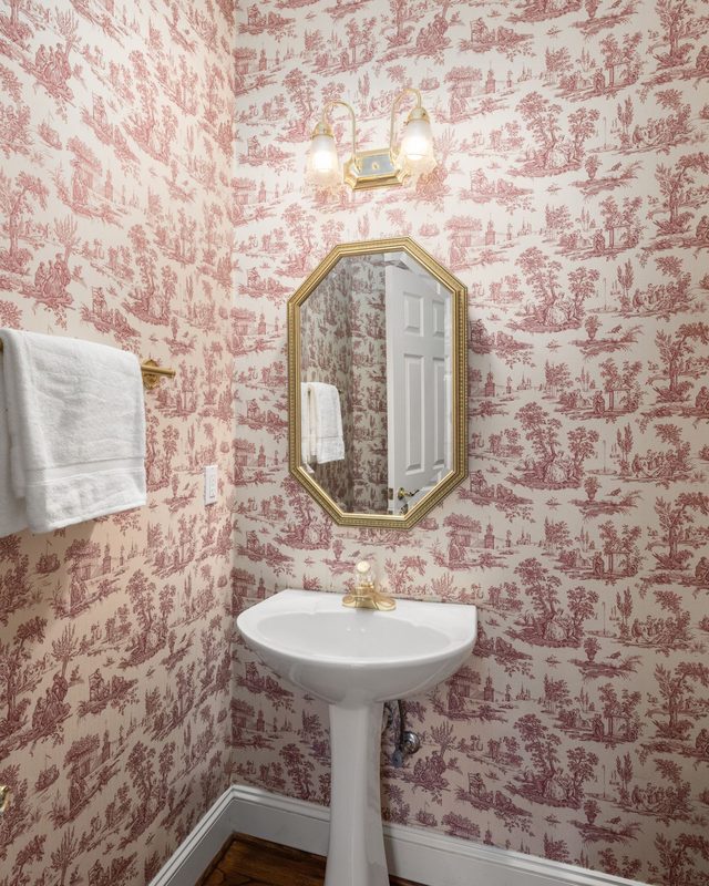 Powder room papered in red-on-cream toile with a pedestal sink and a gold octagonal mirror
