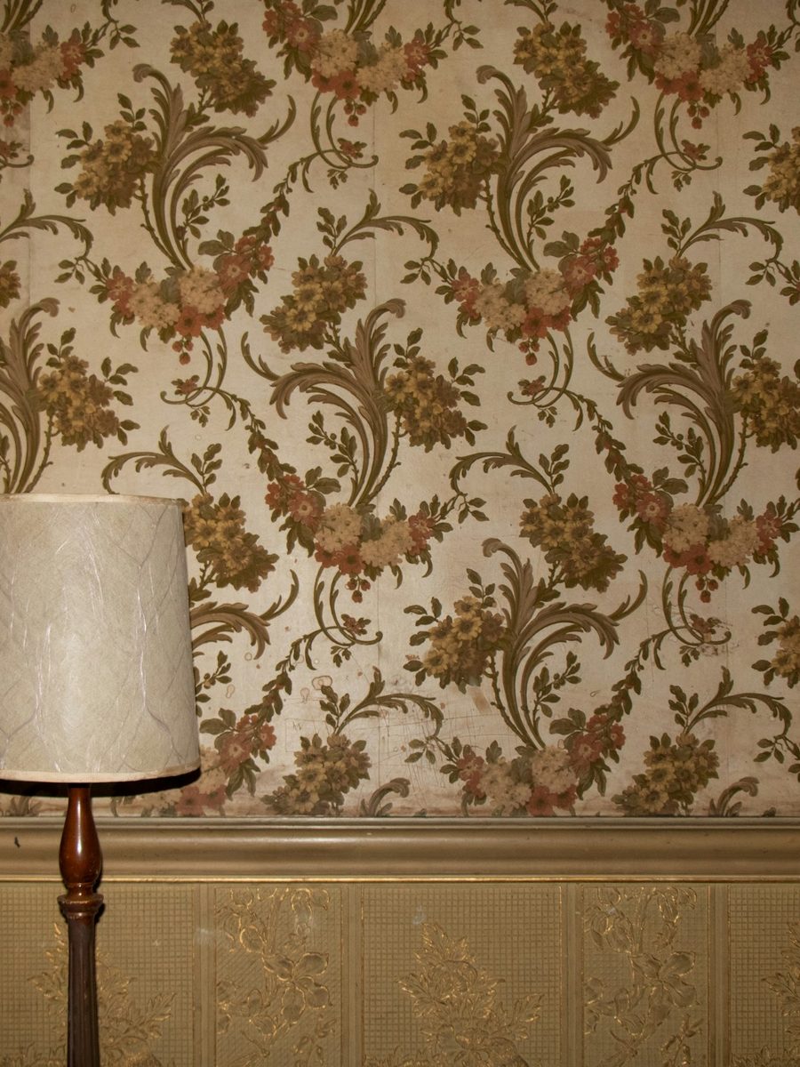 Cream and olive floral wallpaper above a gold-toned dado rail, lit by a floor lamp with a textured shade